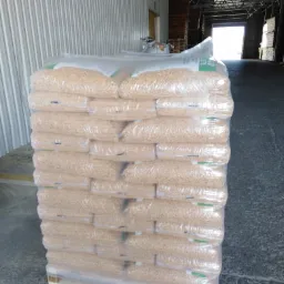 Stos worków z pelletem na palecie, zabezpieczonych folią, w magazynie z metalowymi ścianami i otwartą bramą w tle.