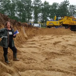 Kobieta w kaloszach i kurtce przeciwdeszczowej z dokumentami na placu budowy z wykopem i ciężkim sprzętem w tle.