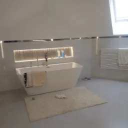 Nowoczesna łazienka na poddaszu z wolnostojącą białą wanną, grzejnikiem drabinkowym z ręcznikiem, oknem dachowym i beżowym dywanikiem na szarych płytkach podłogowych.