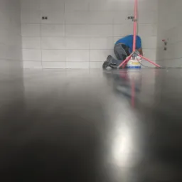 Pracownik na kolanach, wygładza ciemną, błyszczącą posadzkę w pomieszczeniu z białymi, prostokątnymi płytkami na ścianach. Widoczny kabel i puszka z preparatem.