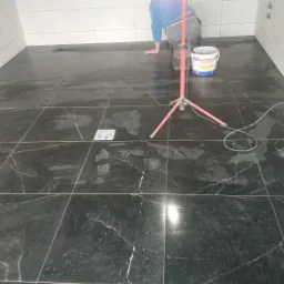 Układanie czarnych płytek imitujących marmur w łazience, widoczny pracownik na kolanach, wiadro z zaprawą, lampa robocza, ślady stóp na świeżo położonych płytkach i gniazdko elektryczne...