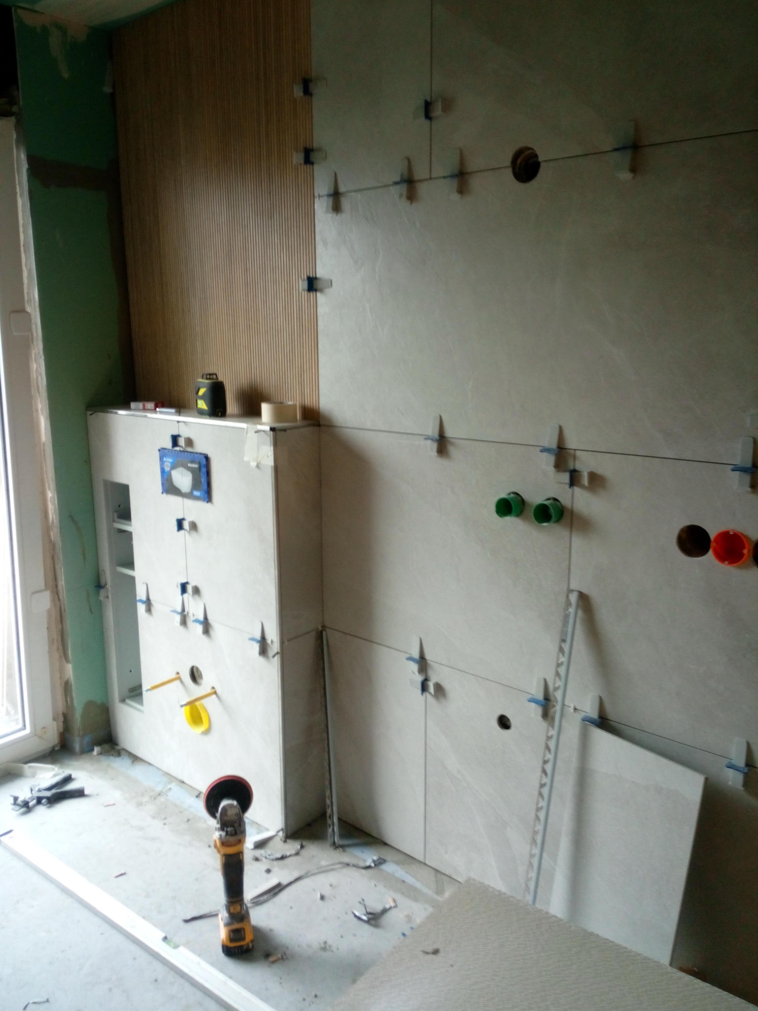 Montaż płytek ściennych w łazience z widocznymi elementami instalacji wodnej i elektrycznej, poziomica laserowa na szafce pod umywalkę, szlifierka kątowa na podłodze, listwa dekoracyjna przy ścianie.