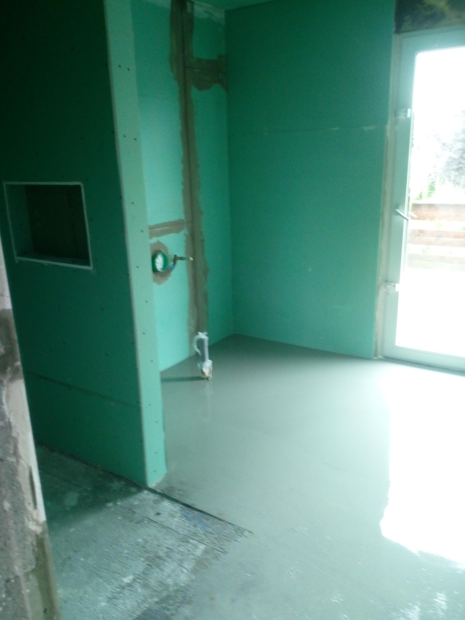 Świeżo wylana, samopoziomująca wylewka na podłodze w łazience z zamontowaną płytą kartonowo-gipsową na ścianach, widoczne przyłącza wodne i odpływ kanalizacyjny, przygotowane do dalszych prac...