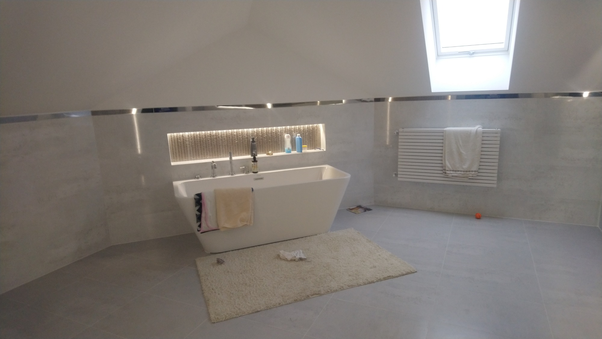 Nowoczesna łazienka na poddaszu z wolnostojącą białą wanną, grzejnikiem drabinkowym z ręcznikiem, oknem dachowym i beżowym dywanikiem na szarych płytkach podłogowych.