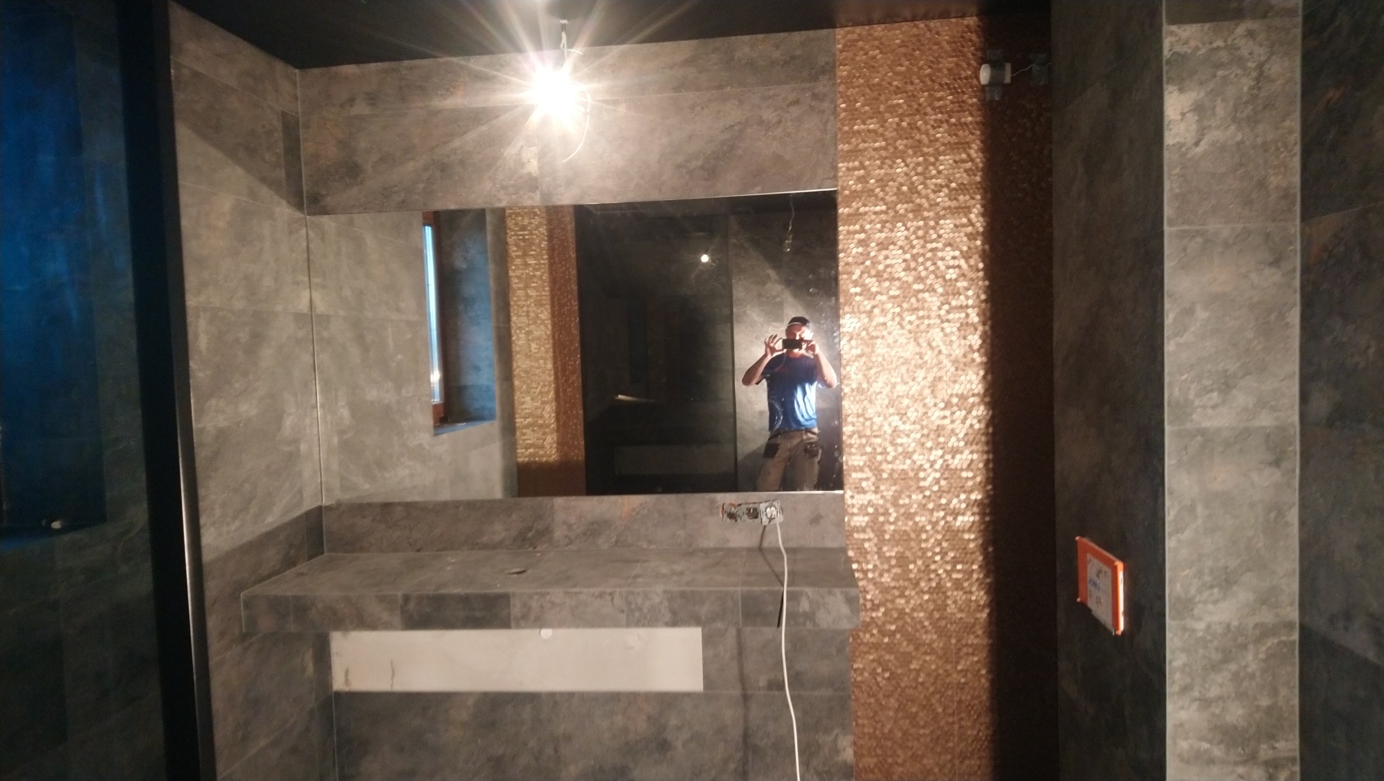 Szara łazienka z lustrem, w którym odbija się osoba robiąca zdjęcie. Na ścianie po prawej stronie złota mozaika, po lewej szare płytki imitujące kamień. Widoczne gniazdka elektryczne...