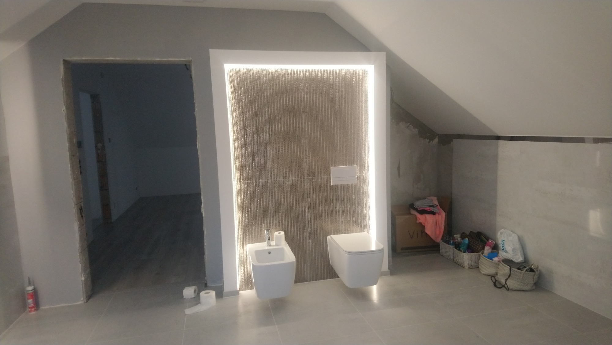 Ukończona łazienka na poddaszu ze skośnym sufitem, widoczne białe sanitariaty: bidet i toaleta podwieszana na tle ściany z dekoracyjnej mozaiki oświetlonej ledami, obok niedokończona ściana...