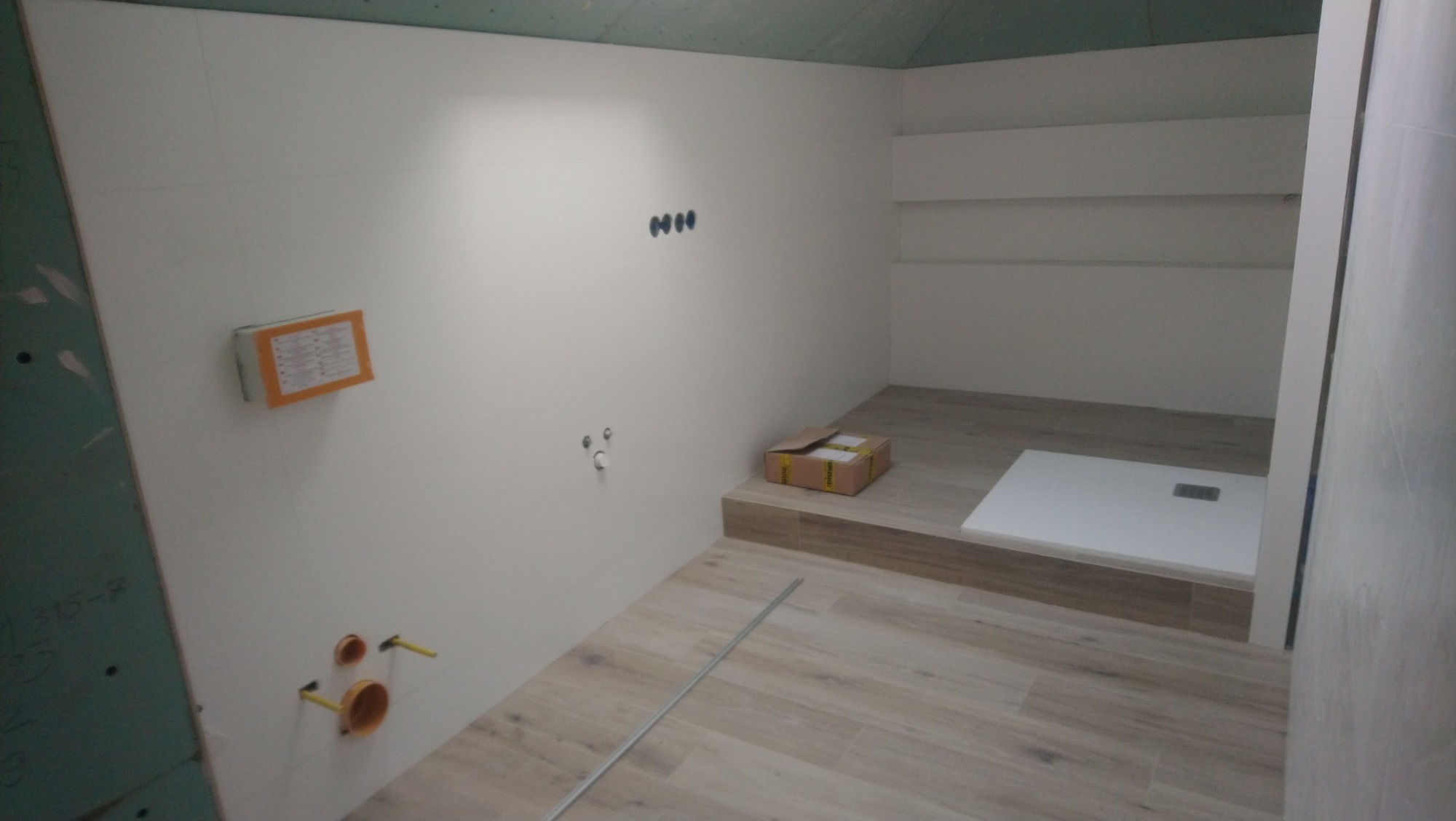 Wnętrze łazienki w trakcie prac wykończeniowych: widoczna kabina prysznicowa, przygotowane instalacje wodno-kanalizacyjne, częściowo położone płytki podłogowe imitujące drewno oraz płyta...