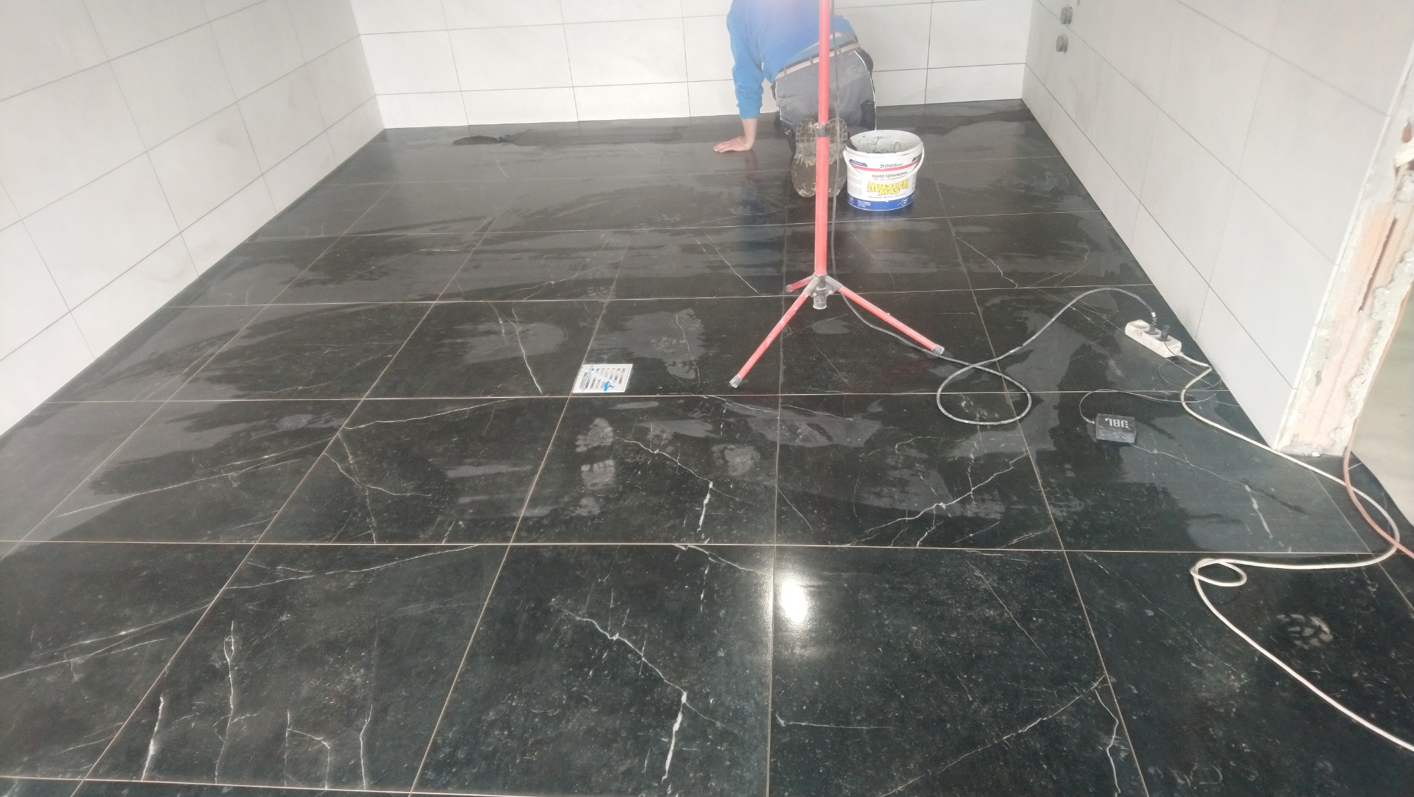 Układanie czarnych płytek imitujących marmur w łazience, widoczny pracownik na kolanach, wiadro z zaprawą, lampa robocza, ślady stóp na świeżo położonych płytkach i gniazdko elektryczne...