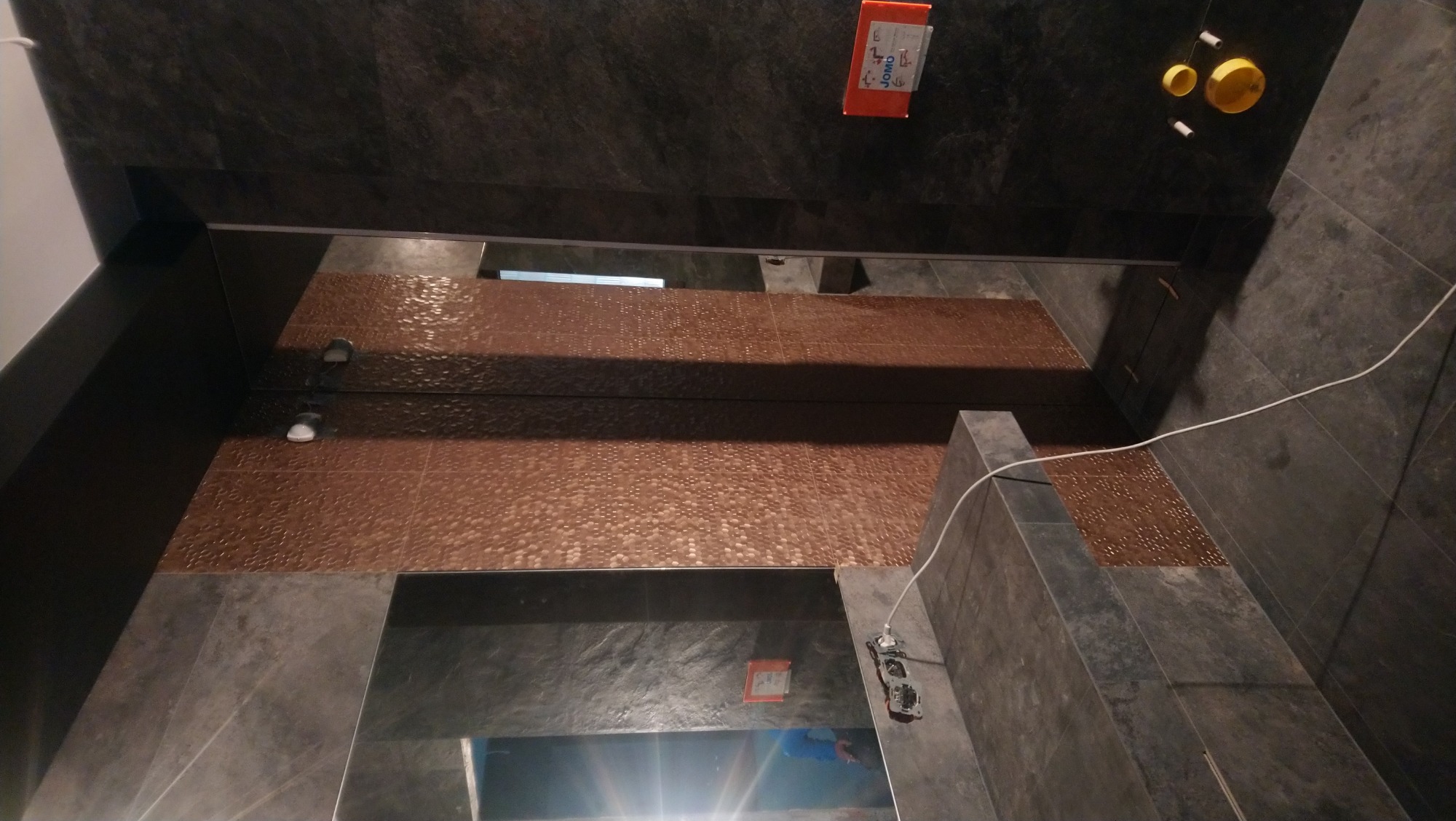 Ciemnoszara łazienka z akcentami miedzianej mozaiki, widok z góry na płytki ścienne i podłogowe z widocznymi elementami instalacji elektrycznej i oświetlenia.