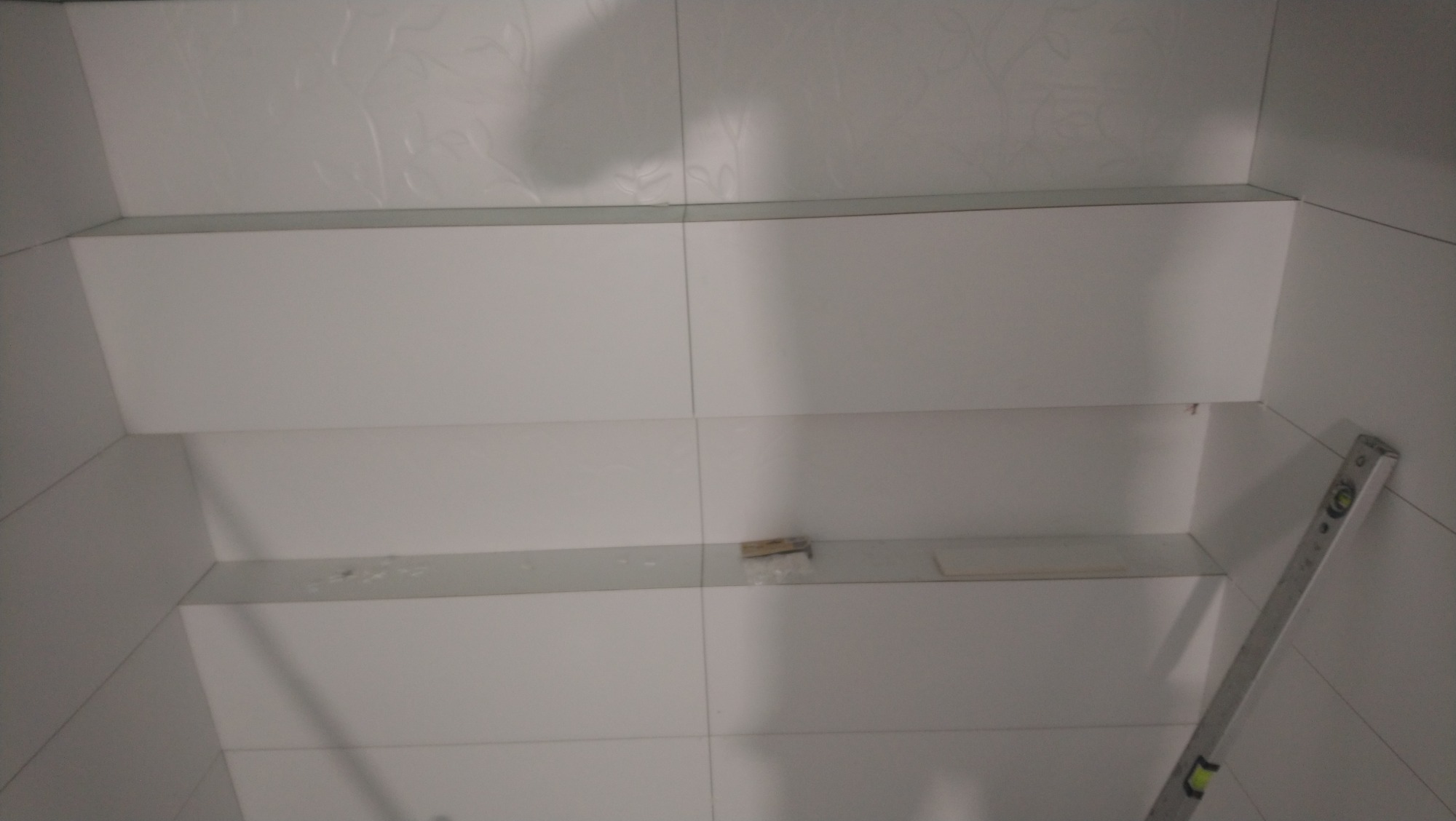 Ściana wyłożona białymi płytkami z półkami w trakcie montażu, widoczna poziomica przy krawędzi.