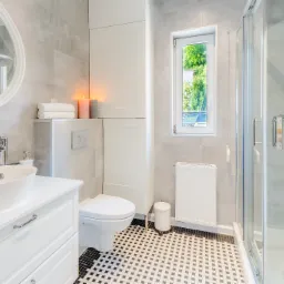 Jasna łazienka z szarymi płytkami, białą armaturą i kabiną prysznicową, akcentowana dekoracyjnymi świecami i czarno-białą mozaiką podłogową.