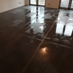 Świeżo położona, ciemna, błyszcząca podłoga z płytek, częściowo mokra, odbijająca światło i okna, widoczne puszki elektryczne w ścianie.