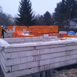 Budowa fundamentów domu z bloczków silikatowych, zabezpieczonych białą plandeką, w tle pomarańczowe opakowania bloczków Silikaty i pracownik w czarnej kurtce.