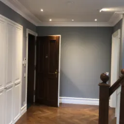 Szare wnętrze domu z drewnianą podłogą w jodełkę, białymi szafami w zabudowie, otwartymi drzwiami z ciemnego drewna i fragmentem drewnianej balustrady schodów.