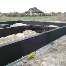 Fundament domu w trakcie budowy, wykonany z czarnych bloczków, częściowo zasypany piaskiem, z widocznym domem w tle.