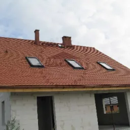 Niedokończony dom murowany z czerwoną dachówką typu karpiówka, trzema oknami dachowymi i dwoma kominami, widoczne ściany z bloczków betonowych.