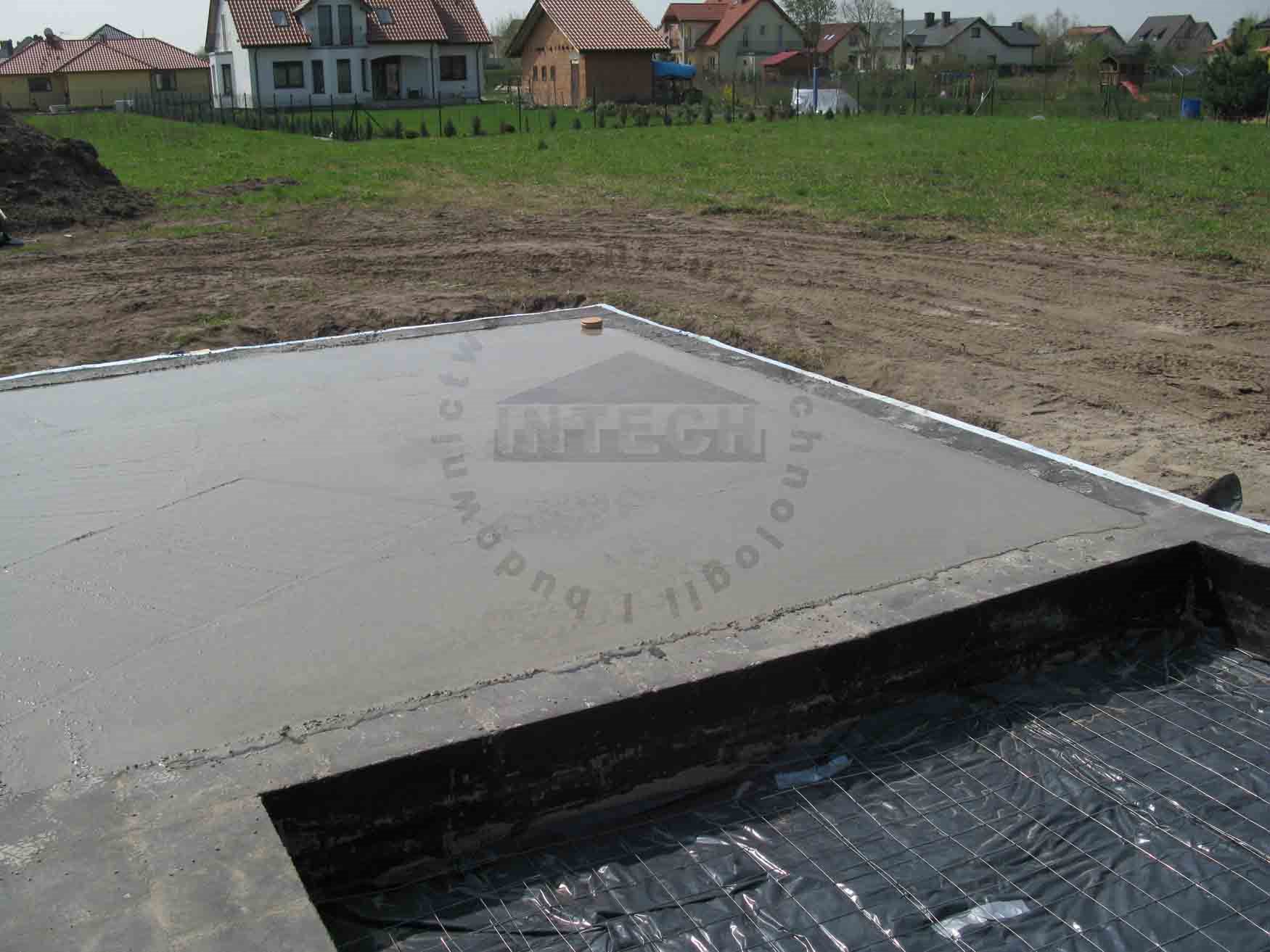 Zalany fundament z widocznym zbrojeniem i folią izolacyjną, w tle zabudowa jednorodzinna.
