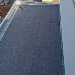 Widok z góry na płaski dach pokryty papą termozgrzewalną, z widocznymi łączeniami i obróbkami blacharskimi przy krawędziach i ścianie budynku.