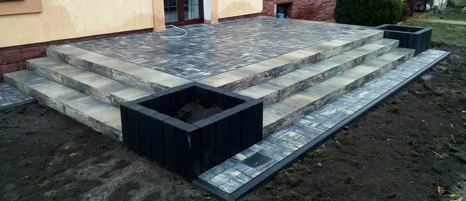 Schody wejściowe z kostki brukowej w odcieniach szarości, zakończone donicami z tego samego materiału, na tle elewacji budynku z widocznym oknem.