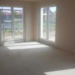 Wnętrze pomieszczenia po nałożeniu tynków maszynowych, widoczne gładkie ściany i sufit, dwa okna z widokiem na zewnątrz, betonowa podłoga z wyraźnymi cieniami słonecznymi.