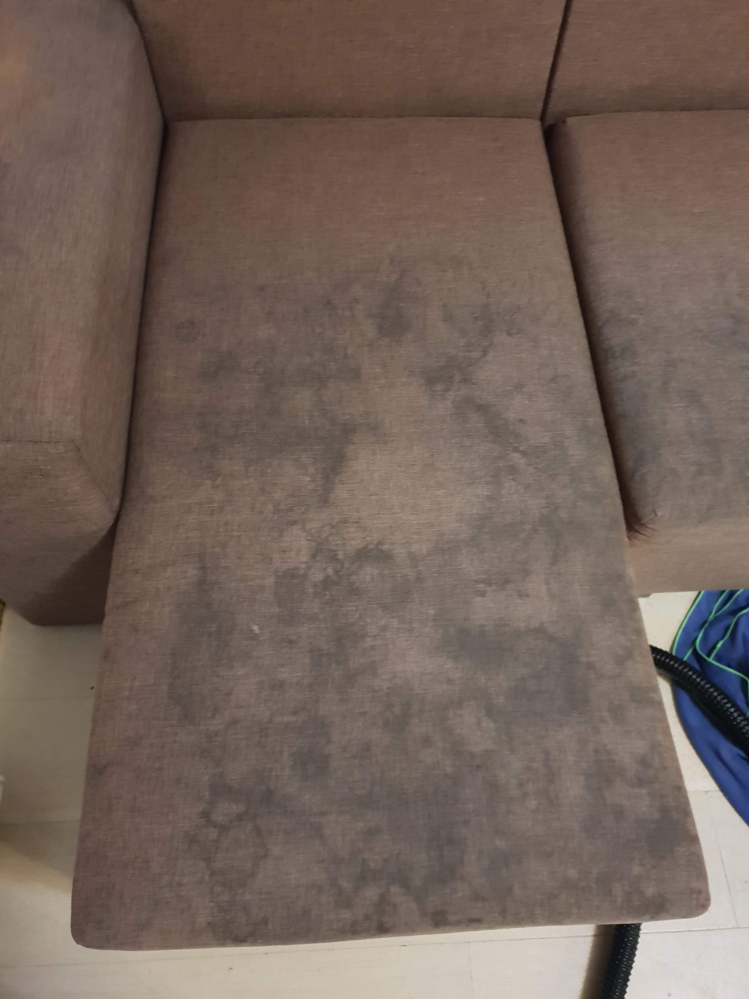 Brązowa kanapa z widocznymi, ciemnymi plamami na siedzisku i oparciu, obok widoczny fragment węża odkurzacza.