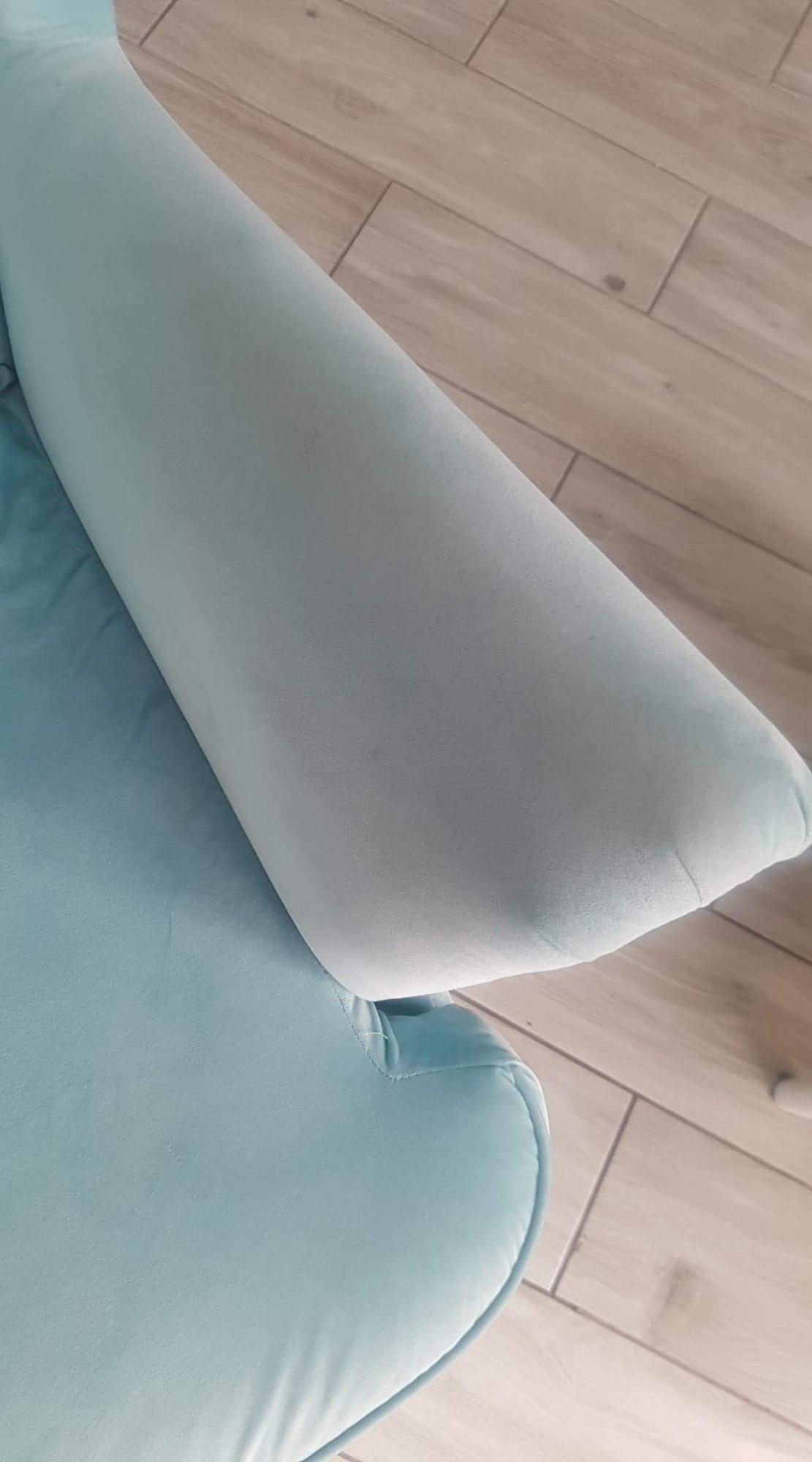 Jasnoniebieski fragment kanapy z widocznym podłokietnikiem i fragmentem siedziska na tle paneli podłogowych imitujących drewno.