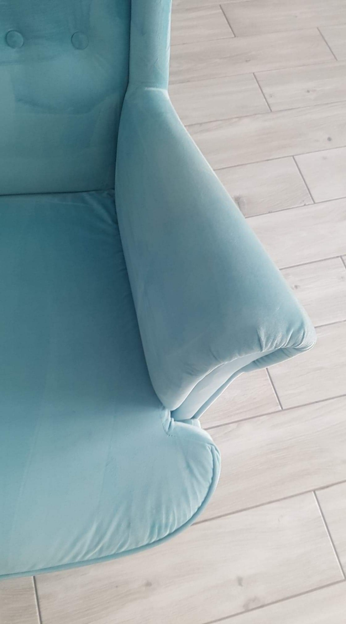 Fragment turkusowego fotela z welurową tapicerką na tle jasnych paneli podłogowych, widoczne detale szwu i delikatne cieniowanie materiału.