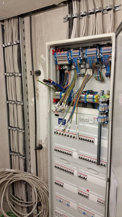 Otwarta skrzynka rozdzielcza z podłączonymi przewodami, widoczne zabezpieczenia i listwy zaciskowe, kable ułożone w korytach kablowych na ścianie w surowym stanie.