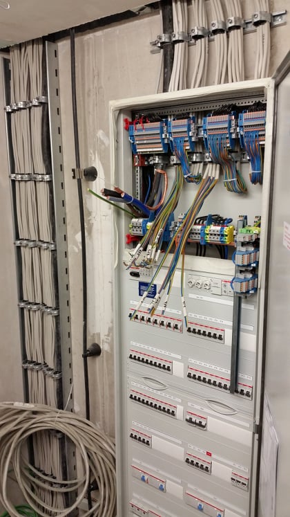 Otwarta skrzynka rozdzielcza z widocznymi bezpiecznikami, przewodami oznaczonymi etykietami i ułożonymi w korytach na ścianie w surowym stanie.