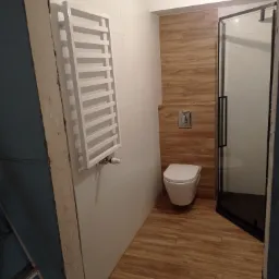 Wykończona łazienka z białymi płytkami, drewnopodobną zabudową WC, grzejnikiem drabinkowym i kabiną prysznicową z czarnymi profilami, sfotografowana z perspektywy niedokończonego wejścia.