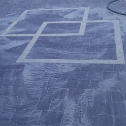 Ułożona, mokra kostka brukowa w odcieniach szarości z dekoracyjnym wzorem z jasnego grysu w kształcie połączonych prostokątów, zielony wąż ogrodowy i metalowa brama w tle.