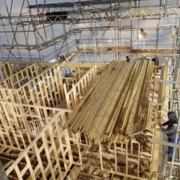 Konstrukcja domu szkieletowego w trakcie budowy, widoczne rusztowania, ułożone drewniane belki i pracownik przycinający drewno na wysokości.