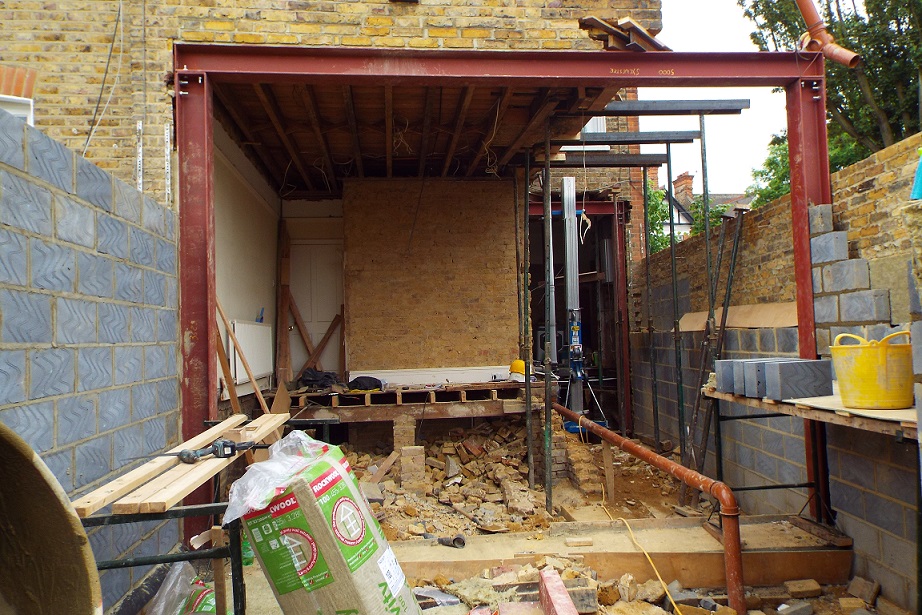 Rozbudowa domu: widok z zewnątrz na konstrukcję stalową, częściowo zdemontowaną podłogę i ściany z cegły oraz bloczków betonowych, z widocznymi materiałami izolacyjnymi Rockwool i narzędziami.