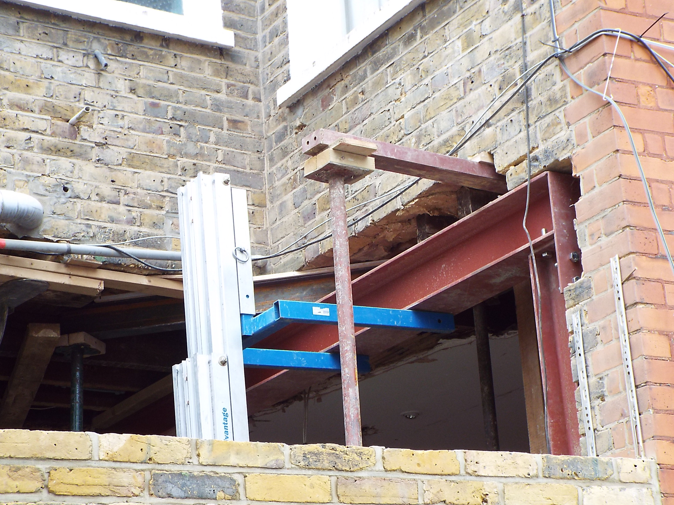 Montaż stalowej belki w murze z cegły podczas remontu, widoczne podpory, drabina aluminiowa i instalacje.