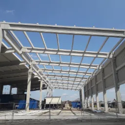 Surowa konstrukcja hali z prefabrykatów betonowych, widok na szkielet dachu i słupy nośne na tle błękitnego nieba, teren budowy zabezpieczony siatką.