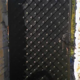 Czarna, łukowata brama z metalu, z dekoracyjnym wzorem rombów, zamontowana między kamiennymi słupkami.