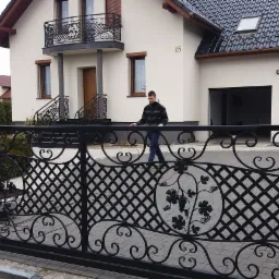 Czarna, kuta brama wjazdowa z motywem winogron, zamontowana przy kamiennym słupku i nowoczesnym domu z balkonem z kutą balustradą, osoba przechodzi w tle.