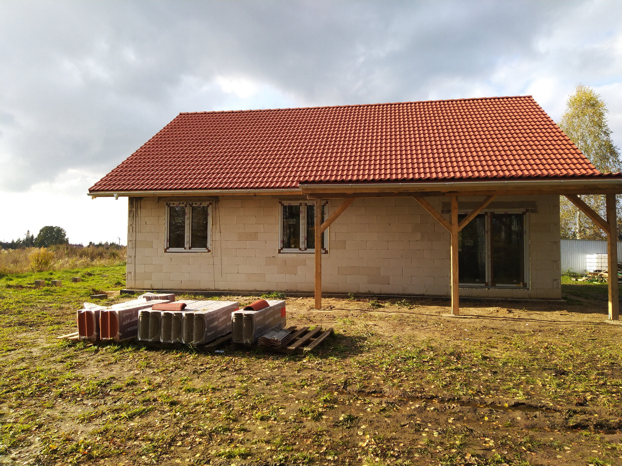 Surowy stan domu jednorodzinnego z czerwonym dachem z dachówki ceramicznej, bloczki betonowe ścian i drewniana konstrukcja zadaszenia tarasu, przygotowane paczki dachówek obok budynku.