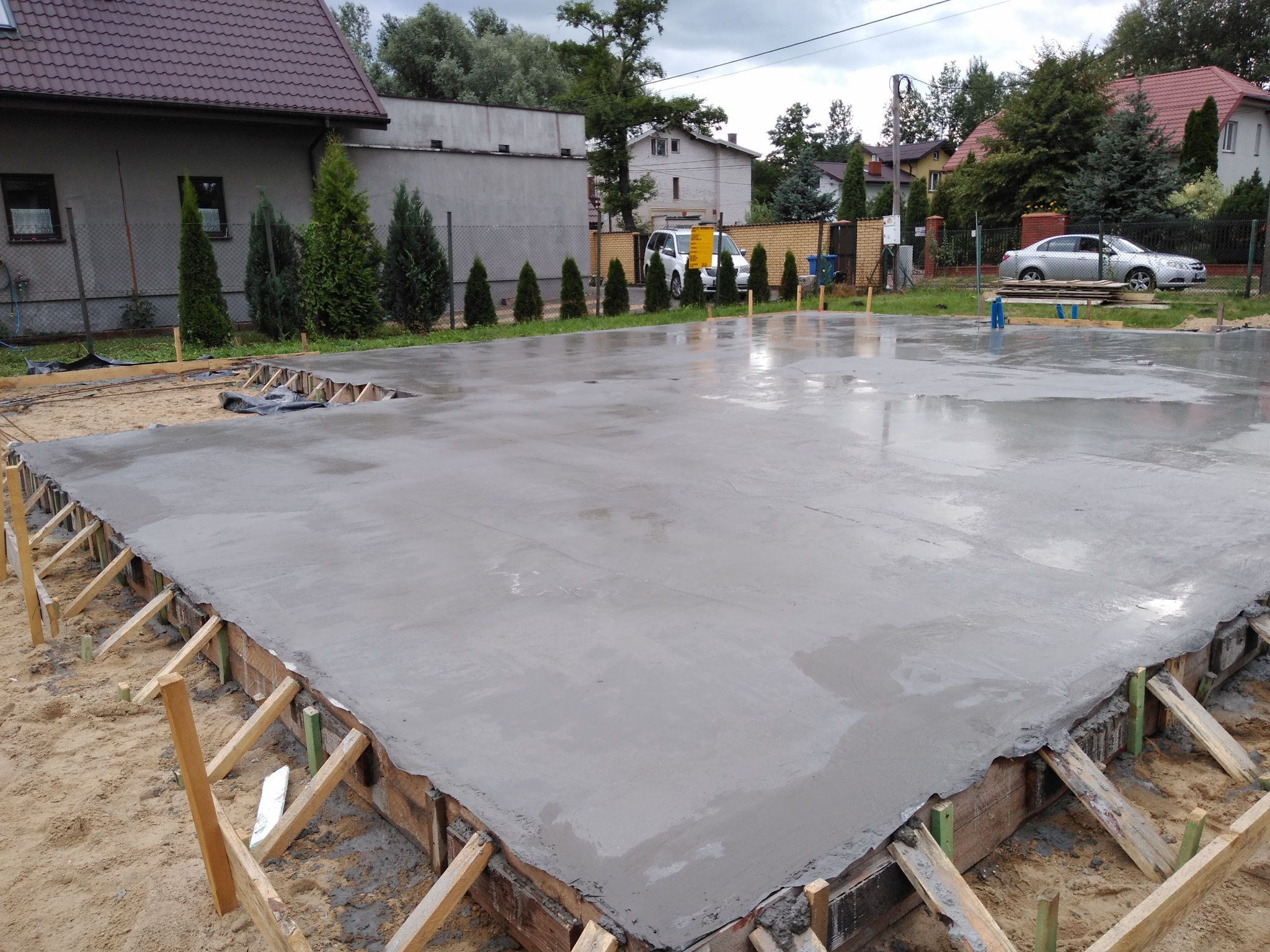 Wylany, mokry beton na przygotowanej płycie fundamentowej z drewnianym szalunkiem na piaszczystym podłożu, widoczne budynki mieszkalne w tle.