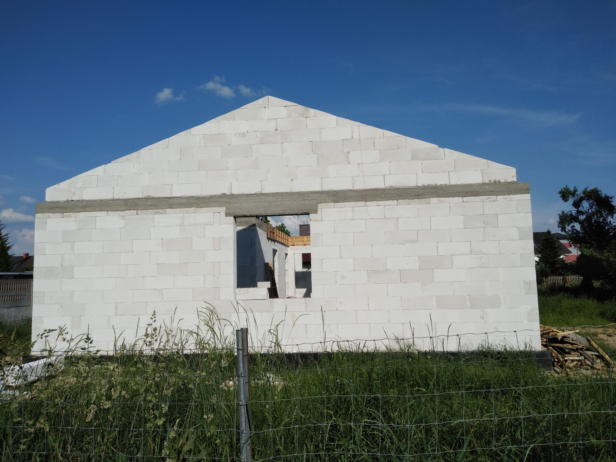 Wznoszone ściany domu z betonu komórkowego, widoczne otwory okienne i fragment drewnianej konstrukcji dachu, na pierwszym planie zarośla i ogrodzenie.