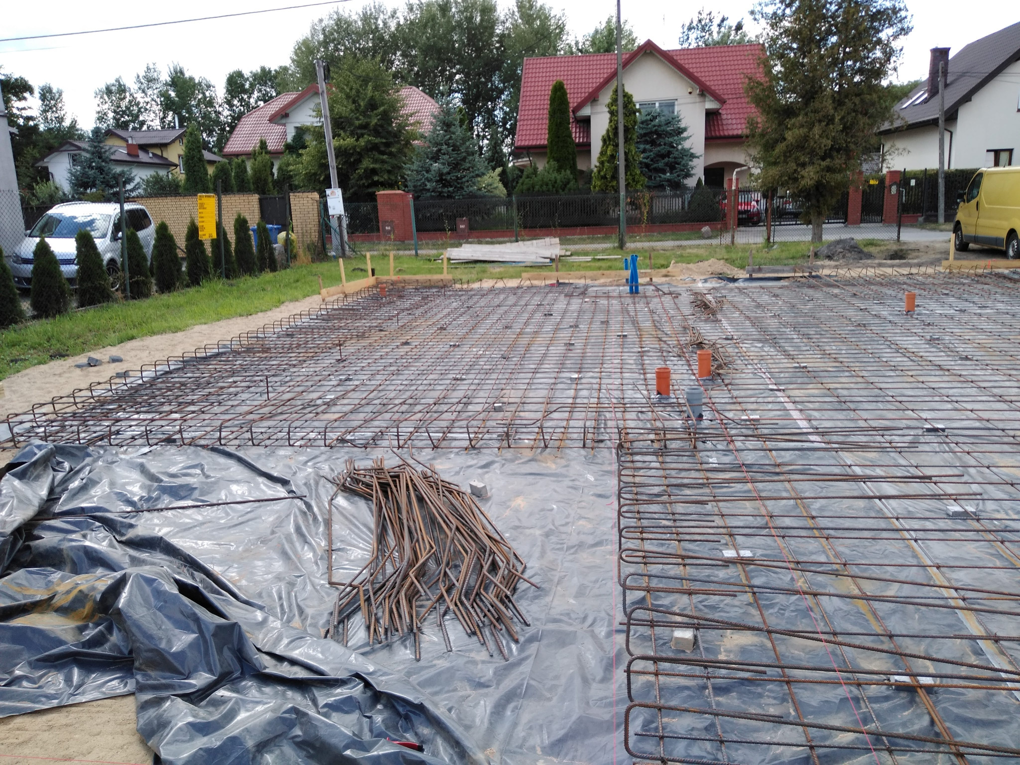 Zbrojenie fundamentów domu jednorodzinnego na folii, widoczne pręty zbrojeniowe i pionowe rury instalacyjne, w tle zabudowa mieszkalna.
