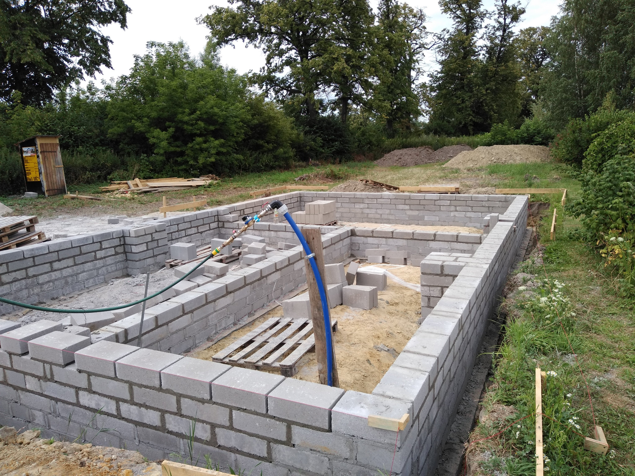 Fundamenty domu w trakcie budowy, ściany z szarych bloczków betonowych, widoczne palety i węże doprowadzające wodę, w tle drzewa i ziemia.