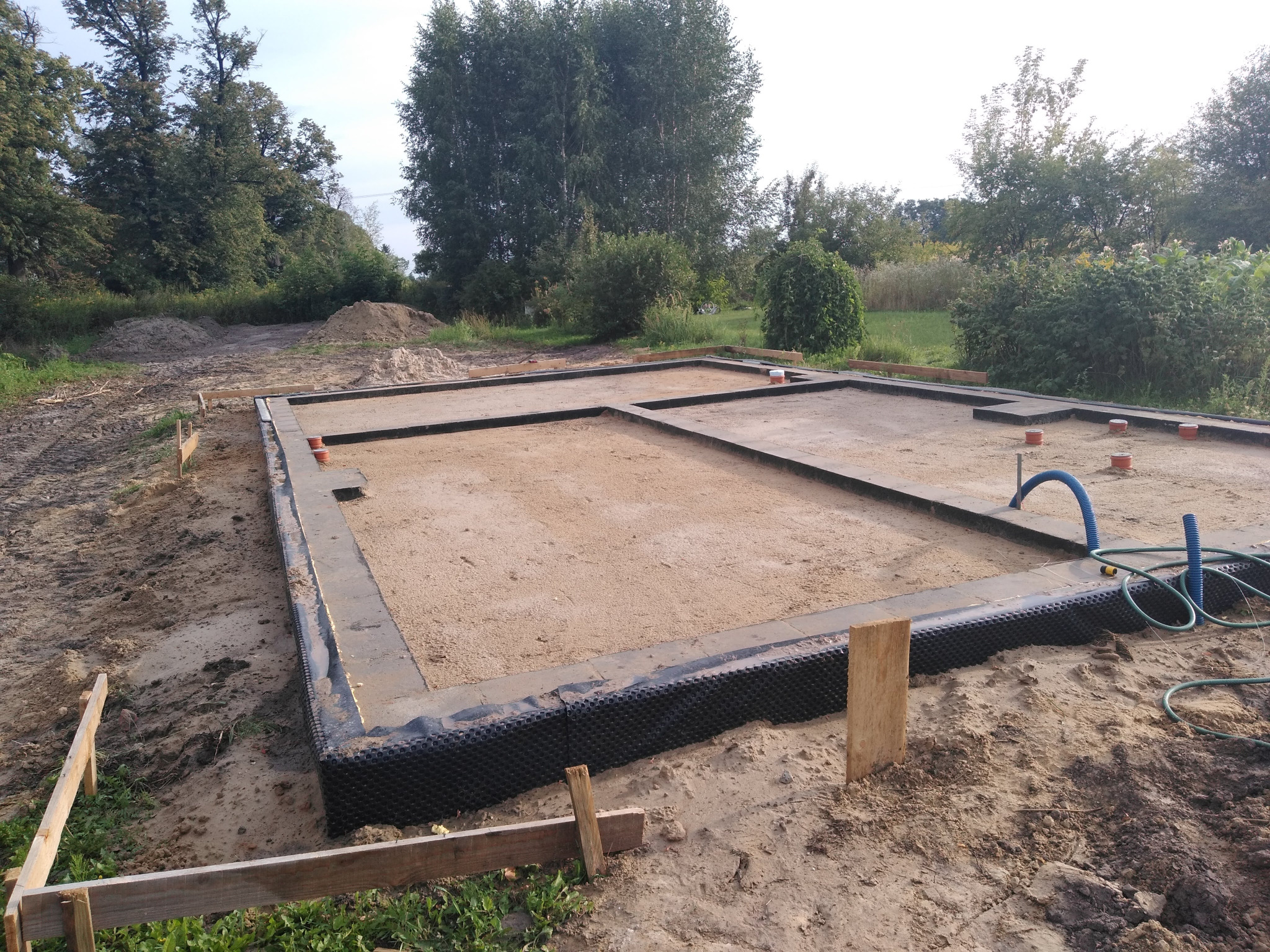 Przygotowany fundament pod budynek jednorodzinny z hydroizolacją pionową, podsypany piaskiem, z widocznymi pionowymi rurami instalacyjnymi i drewnianym szalunkiem.
