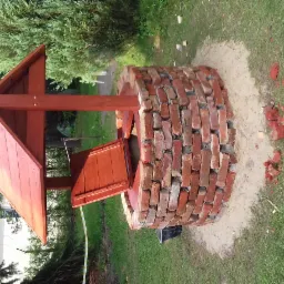 Studnia z czerwonej cegły z drewnianym daszkiem, widoczne gruz i pozostałości po budowie na trawie.