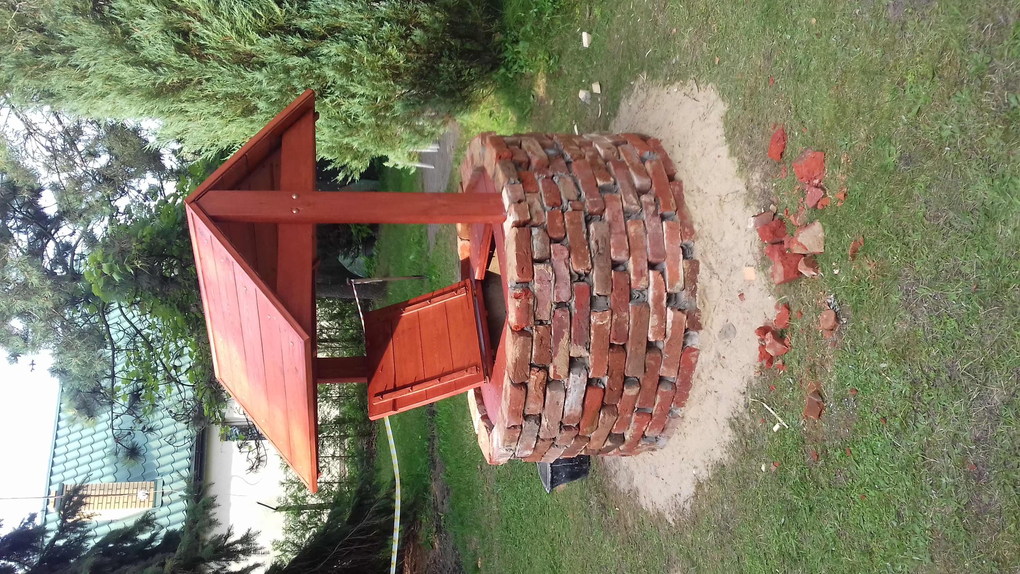 Studnia z czerwonej cegły z drewnianym daszkiem, widoczne gruz i pozostałości po budowie na trawie.