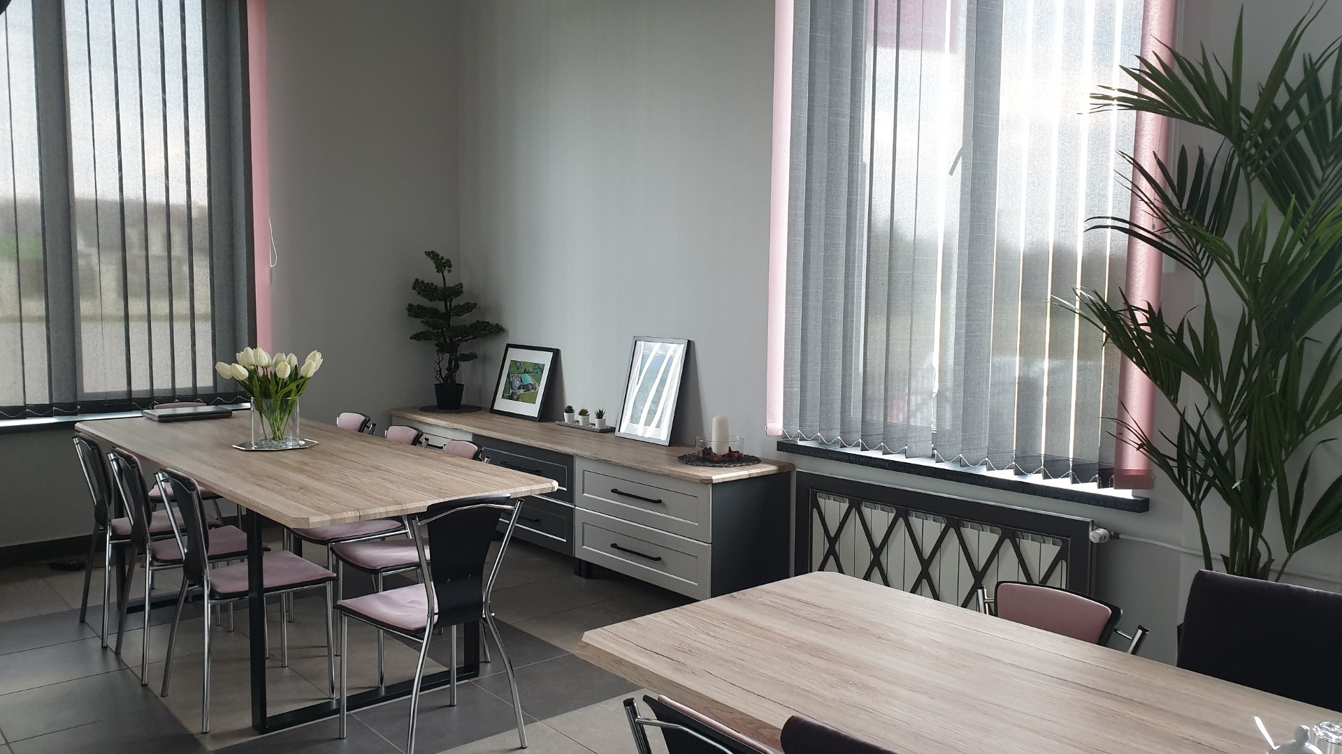 Jasne wnętrze biurowe z dwoma drewnianymi stołami, krzesłami z różowym obiciem, szafkami pod oknami i kwiatami w wazonie na jednym ze stołów.