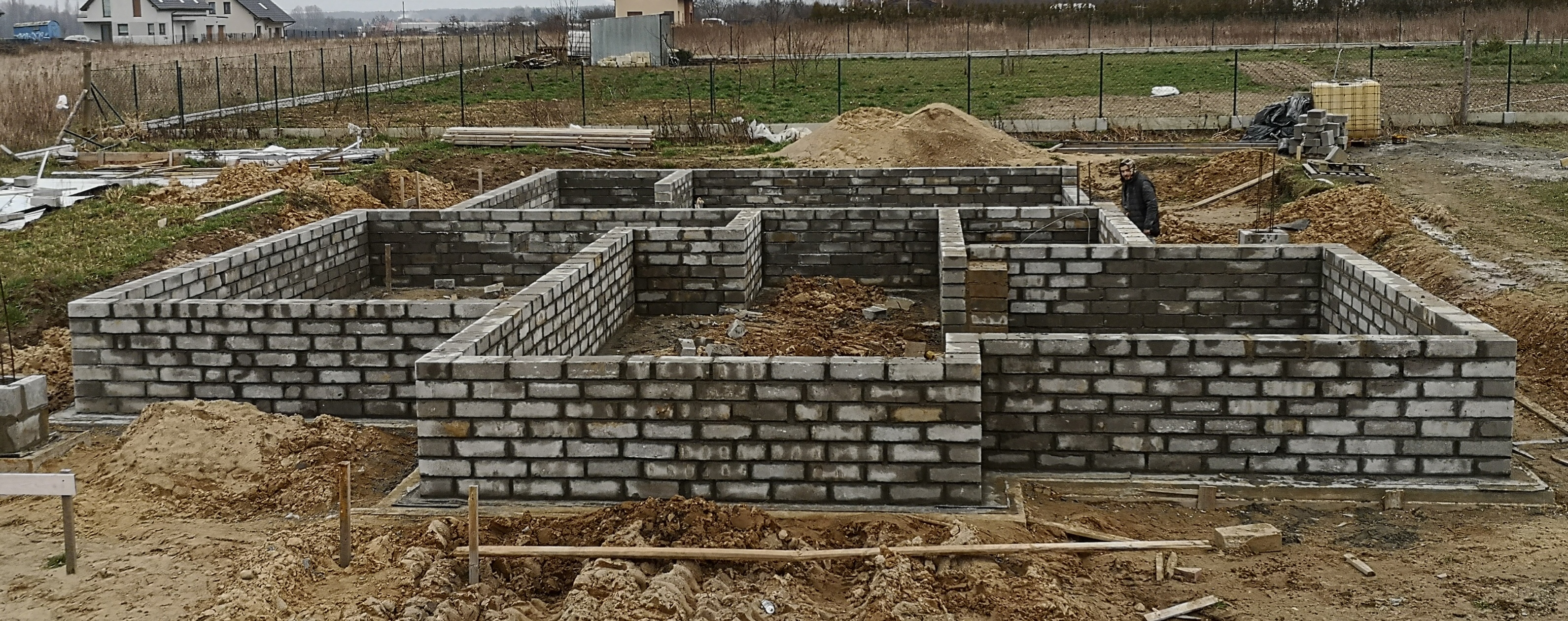 Wznoszenie fundamentów z bloczków betonowych na placu budowy, widoczny zarys przyszłego budynku, ziemia i materiały budowlane wokół.