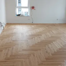 Jasna, pusta przestrzeń z ułożonym parkietem w jodełkę, widoczne niedokończone prace i kable wystające ze ściany, z fragmentem szarych płytek podłogowych na pierwszym planie.