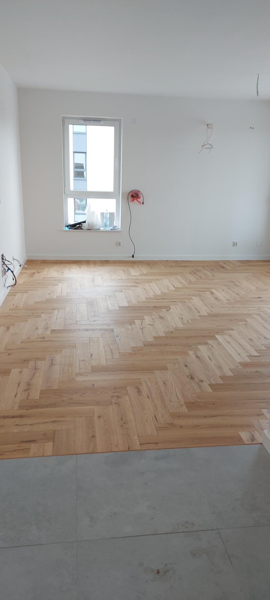 Jasna, pusta przestrzeń z ułożonym parkietem w jodełkę, widoczne niedokończone prace i kable wystające ze ściany, z fragmentem szarych płytek podłogowych na pierwszym planie.