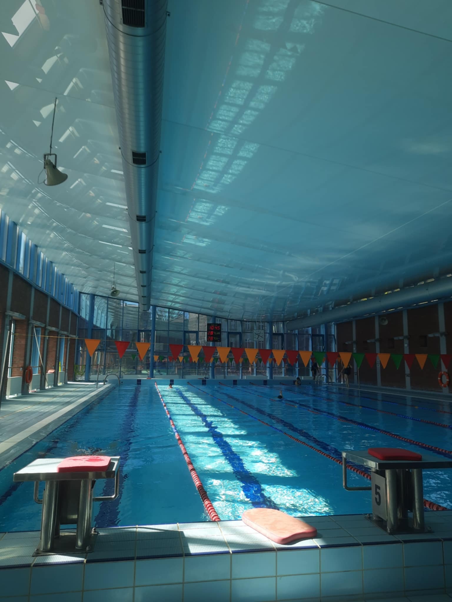 Kryty basen z błękitną wodą, torami oddzielonymi linami, słupkami startowymi i dekoracyjnymi flagami. Widoczne osoby pływające.
